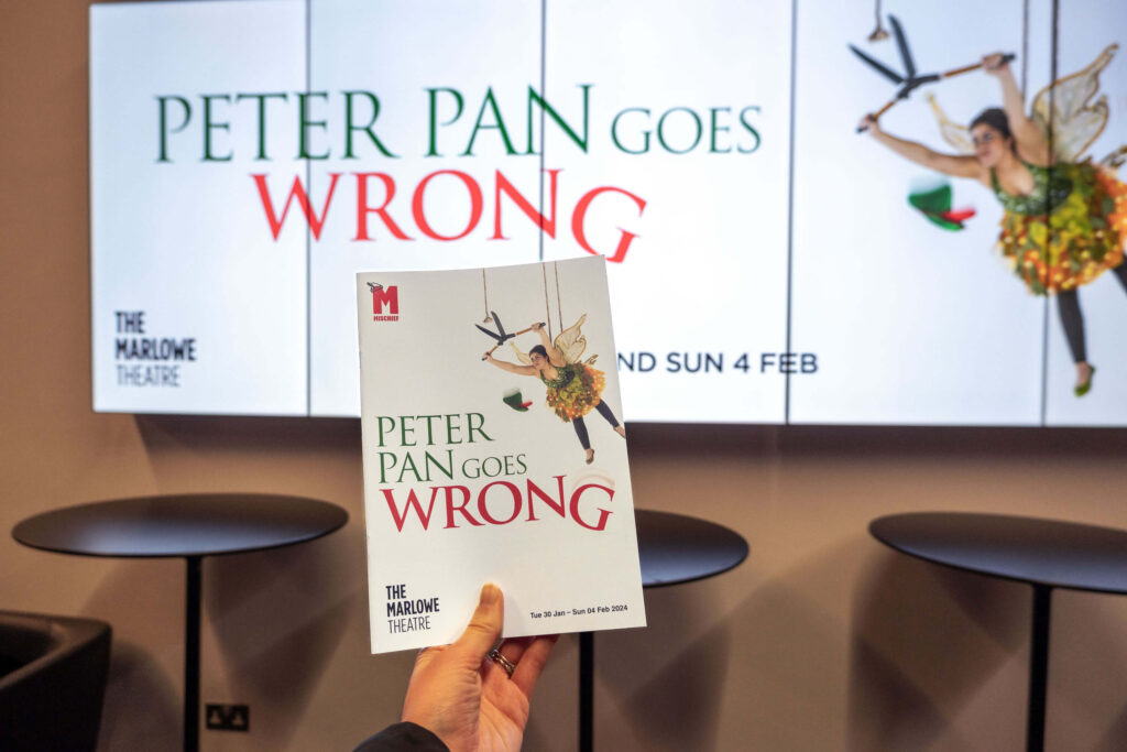 Peter Pan Goes Wrong programme in front of the digital poster at The Marlowe Theatre, Canterbury
