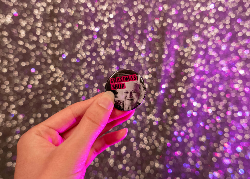 Julie Flower: Grandma's Shop badge featuring Hilda Flower in front of the Gilded Balloon Patter House glitter wall | Edinburgh Fringe